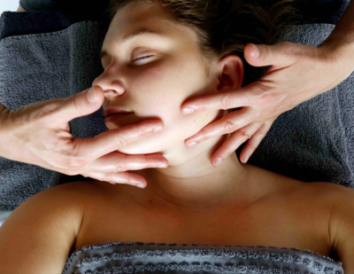 Une jeune femme apprécie son massage Kobido. Elle est couchée sur le dos, la tête légèrement tournée vers la gauche, les cheveux tirés vers l’arrière et son corps est enveloppé par une serviette douillette. La ligne de la mâchoire est ici lissée entre l’index et le majeur des deux mains expérimentées de la praticienne. Le mouvement va du menton vers l’oreille.